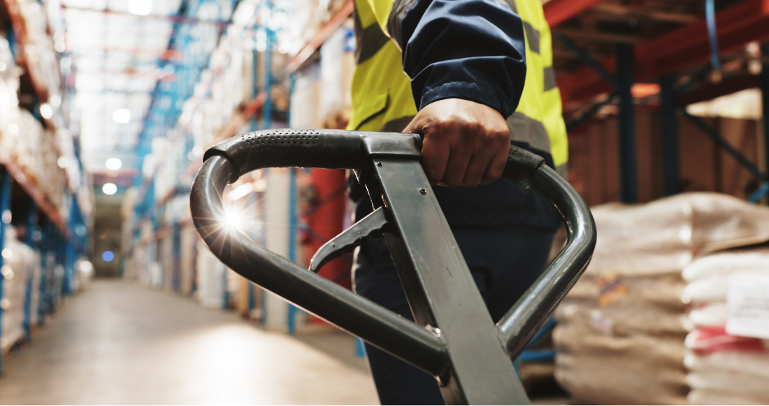 Une personne portant un gilet de haute visibilité pousse un transpalette manuel dans une allée d’un entrepôt, entouré d’étagères remplies de marchandises et de colis divers. Une personne portant un gilet de haute visibilité pousse un transpalette manuel dans une allée d'un entrepôt, entouré d'étagères remplies de marchandises et de colis divers.