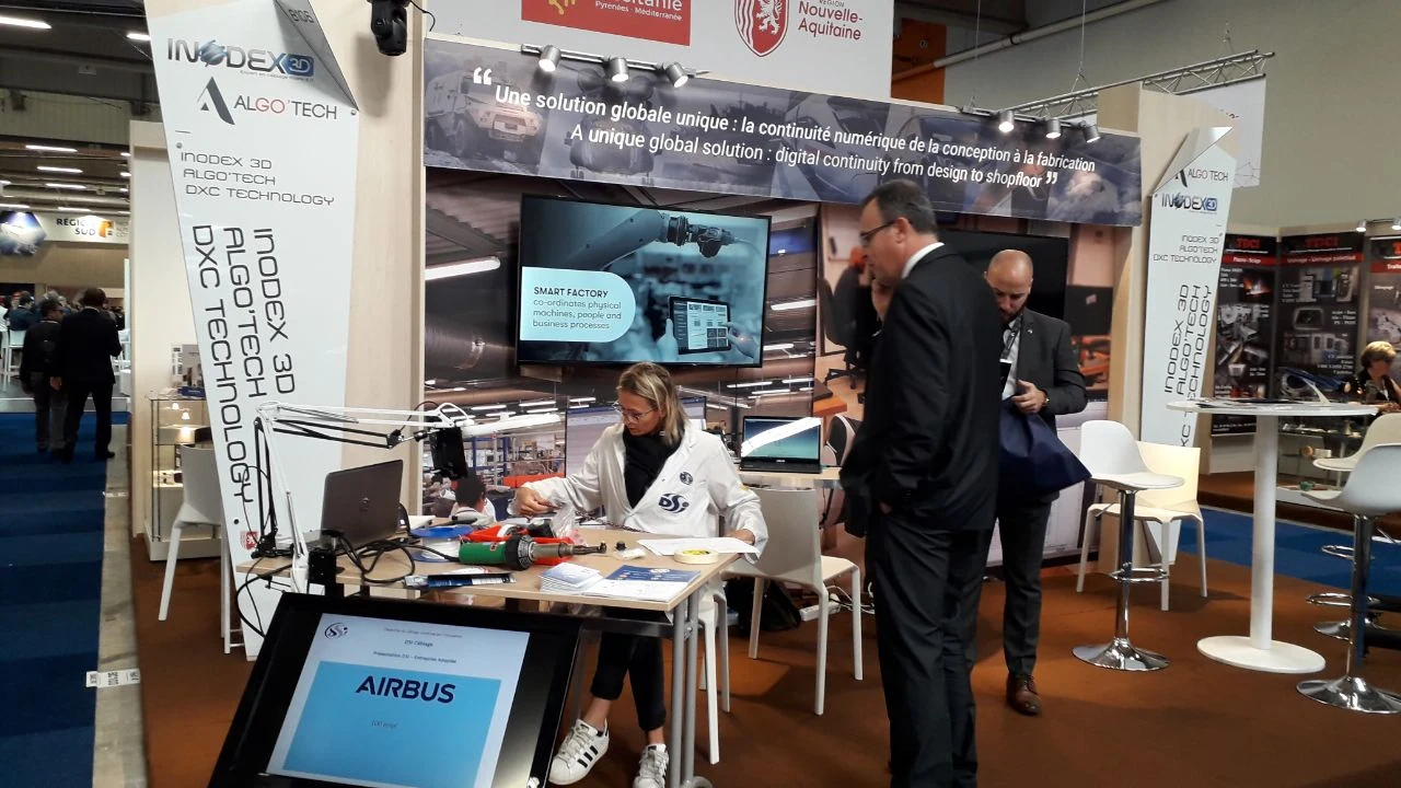 Une femme est assise à une table avec des appareils électroniques et des outils sur un stand d'exposition technologique, tandis que deux hommes en costume se tiennent à proximité. Le stand présente des écrans, des bannières et des panneaux présentant Airbus et des solutions de fabrication numérique.
