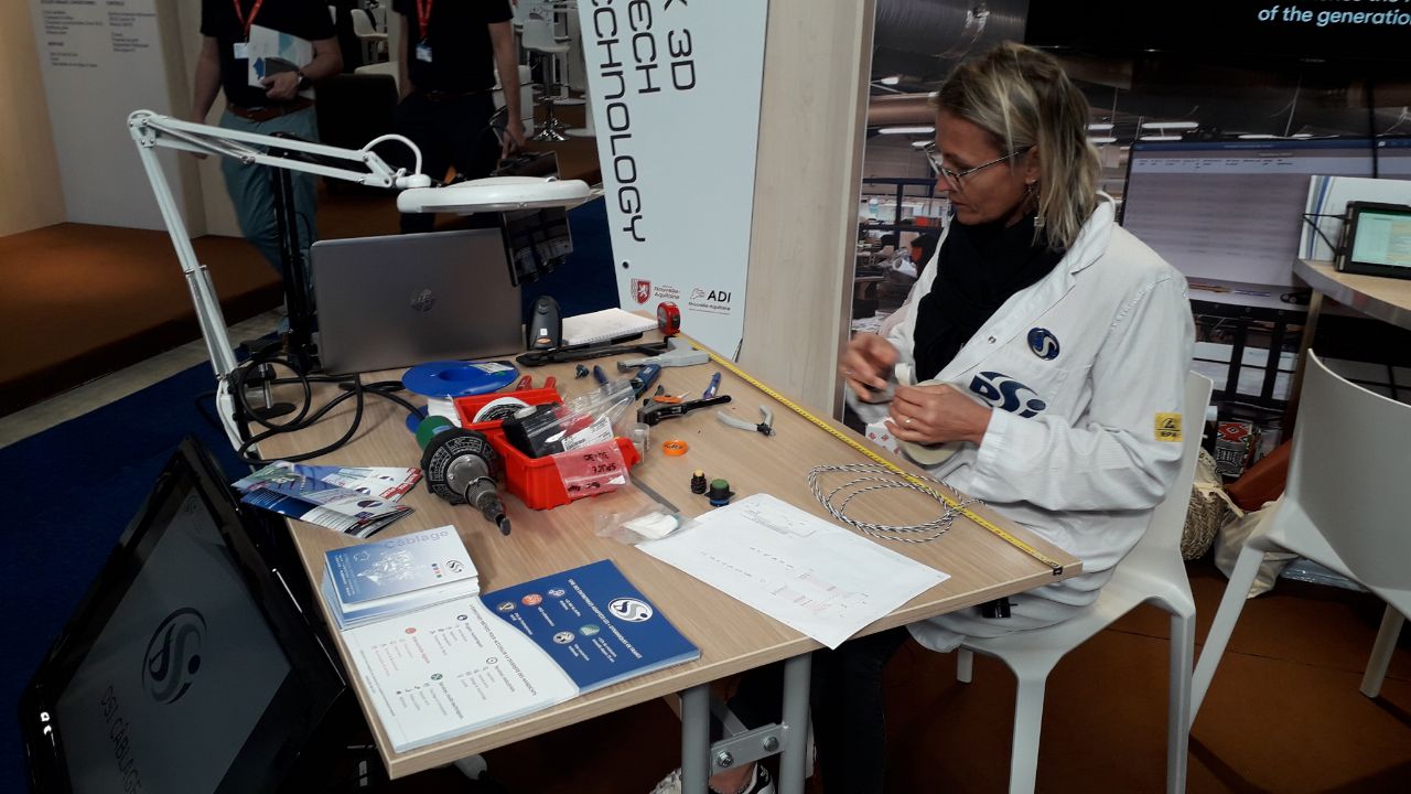 Une femme en blouse blanche est assise à un bureau avec des outils, des appareils électroniques et des documents techniques, travaillant sur un projet lors d'un événement ou d'un atelier technologique. Un ordinateur portable et divers équipements sont posés sur la table.