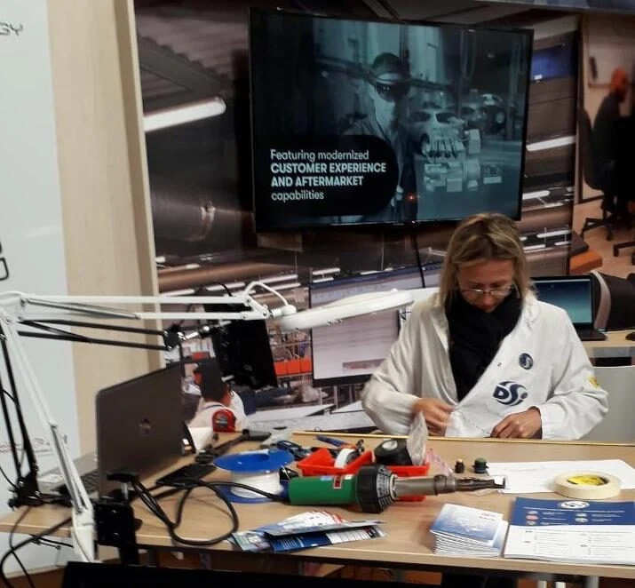 Une femme en blouse blanche travaille sur des composants électroniques à un bureau équipé d'outils. Au premier plan, un écran affiche le logo et le site web de DSI. Un grand écran affichant des images de machines est visible en arrière-plan.