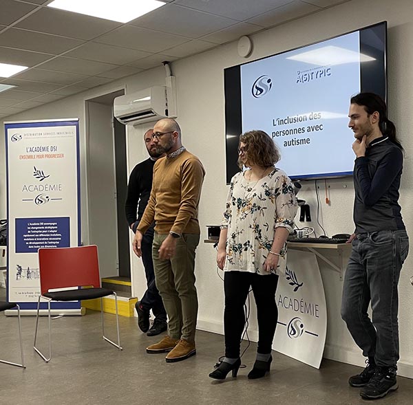 Quatre personnes se tiennent à l'avant d'une salle lors d'une présentation. Derrière elles, un écran affiche le titre L'inclusion des personnes avec autisme et une bannière de l'ACADÉMIE SSi. Deux chaises vides sont visibles au premier plan.