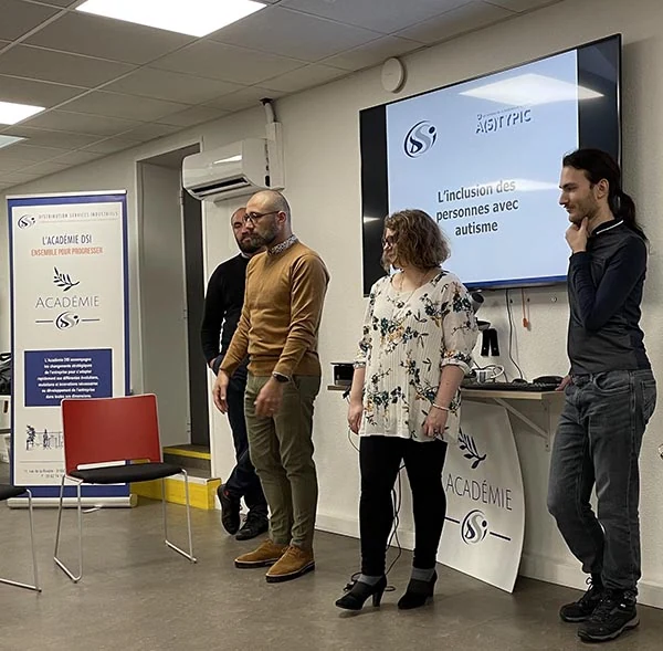 Quatre personnes se tiennent à l'avant d'une salle lors d'une présentation. Derrière elles, un écran affiche le titre L'inclusion des personnes avec autisme et une bannière de l'ACADÉMIE SSi. Deux chaises vides sont visibles au premier plan.