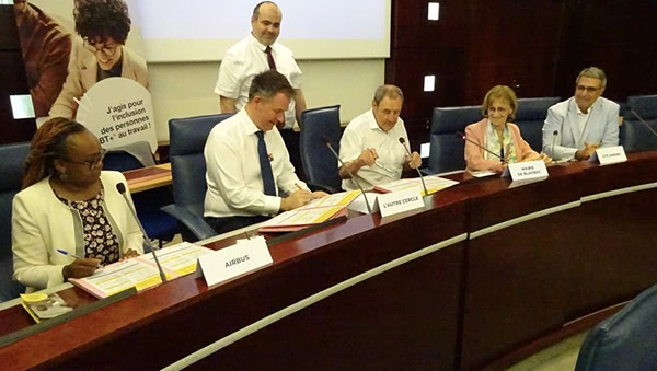 Cinq personnes sont assises à une table de conférence et signent des documents, avec des plaques nominatives devant elles. Un homme se tient derrière eux et une bannière avec du texte et l'image d'une femme souriante est affichée en arrière-plan.