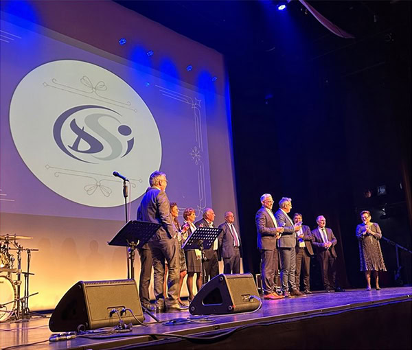 Un groupe de personnes se tient sur une scène sous des projecteurs, avec des microphones et des pupitres à proximité. Un grand logo est projeté sur un écran derrière eux. L'atmosphère suggère une cérémonie de remise de prix ou un événement formel.