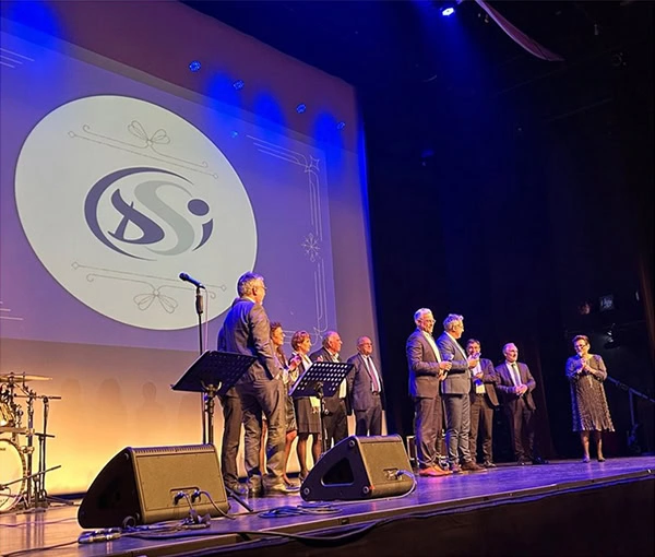 Un groupe de personnes se tient sur une scène sous des projecteurs, avec des microphones et des pupitres à proximité. Un grand logo est projeté sur un écran derrière eux. L'atmosphère suggère une cérémonie de remise de prix ou un événement formel.