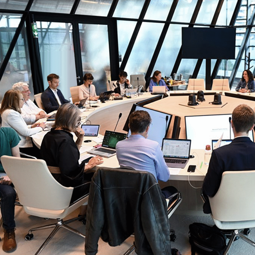 Un groupe de personnes est assis autour d'une grande table de conférence moderne, avec des ordinateurs portables et des documents, et participe à une réunion à l'intérieur d'un bureau aux murs de verre. Un grand écran est visible à l'avant de la salle.