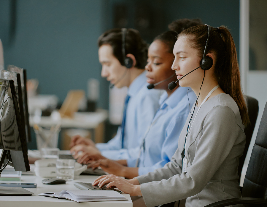 Trois représentants du service client portant des casques sont assis en rangée à des bureaux, travaillant sur des ordinateurs dans un environnement de bureau moderne.