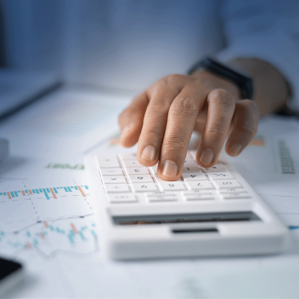 Une main appuie sur les boutons d'une calculatrice blanche, avec des documents commerciaux et des graphiques financiers visibles sur le bureau en arrière-plan.