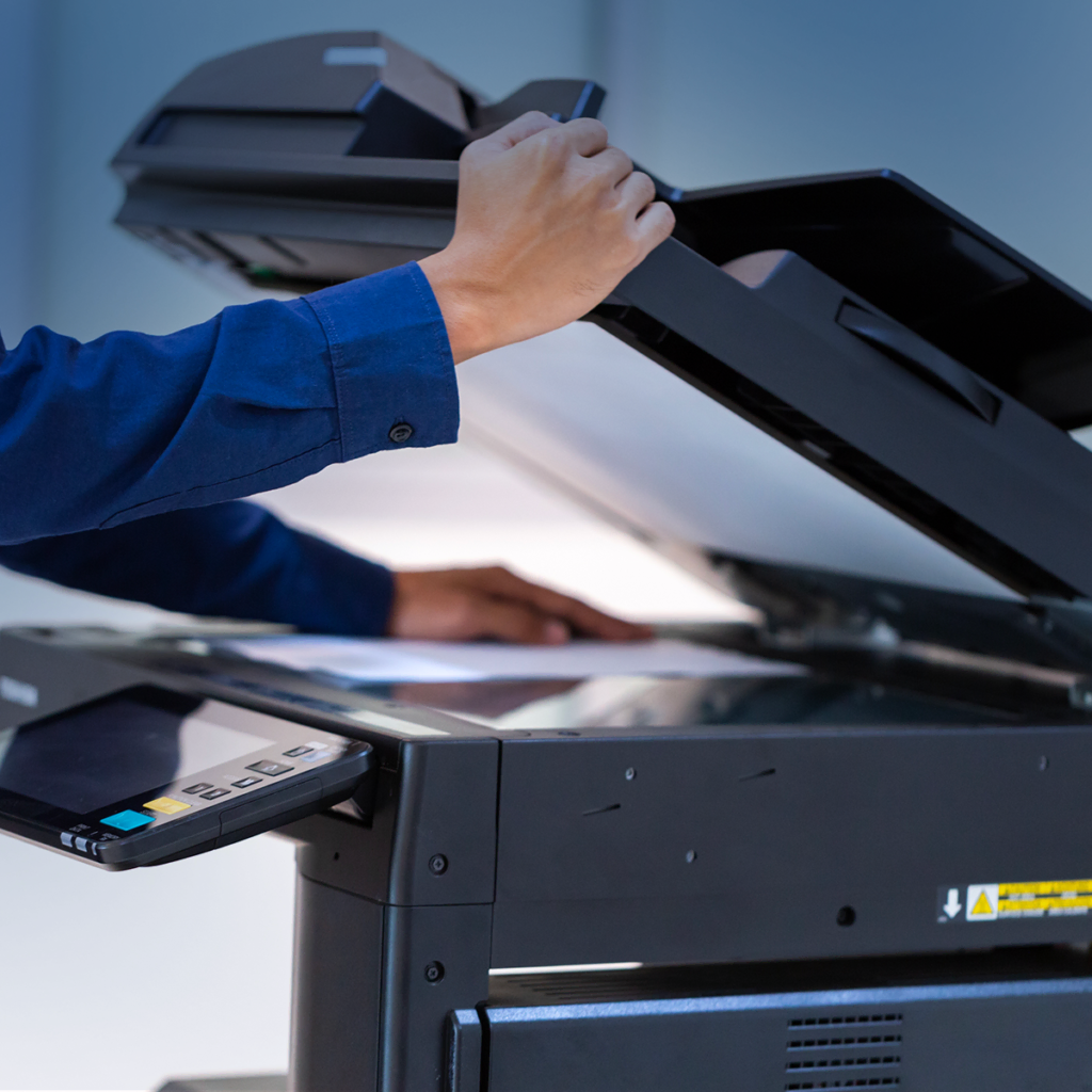 Une personne en chemise bleue soulève le couvercle d'un grand copieur ou scanner de bureau, se préparant à placer ou à retirer un document sur la surface de numérisation en verre.