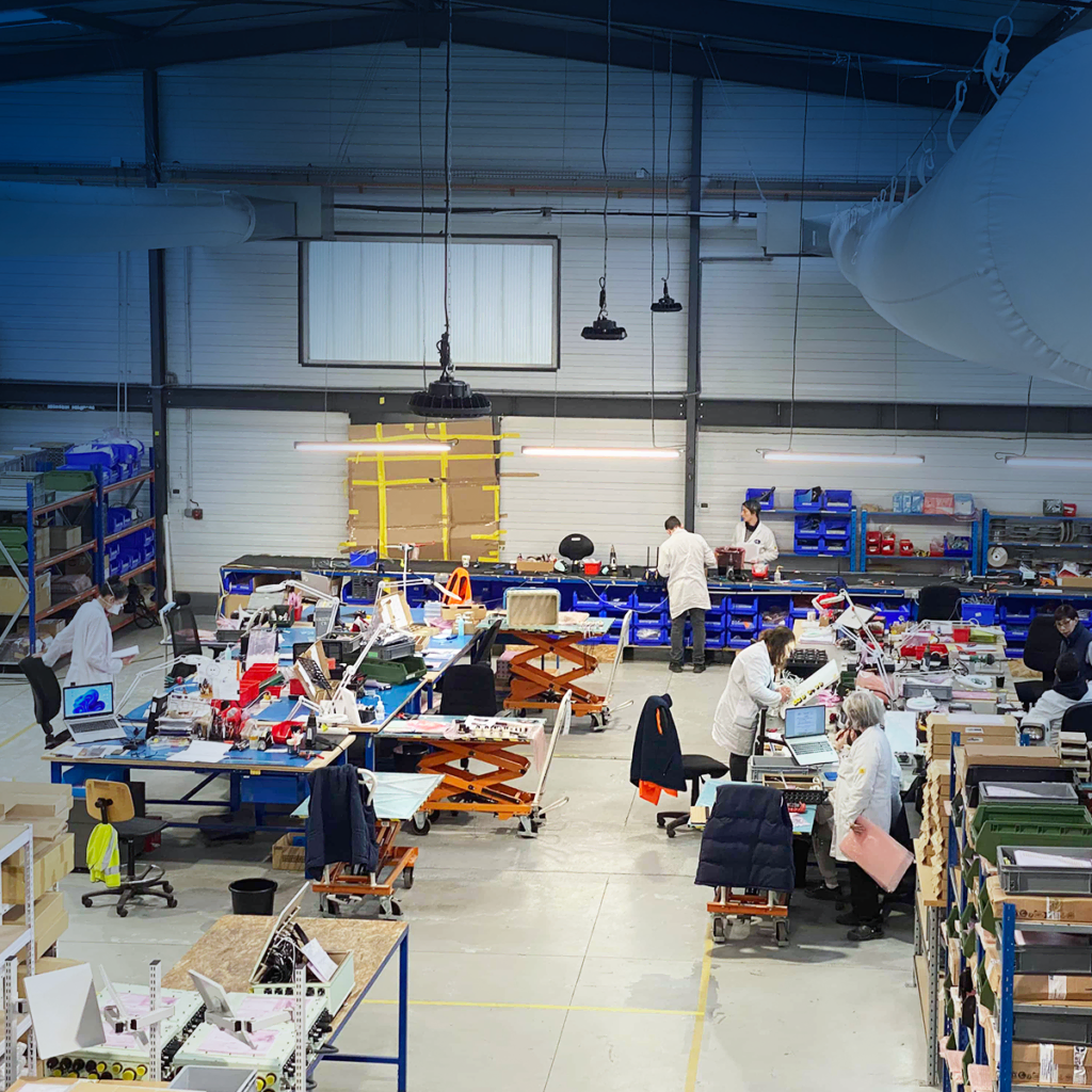Un groupe de personnes travaille à des tables dans un vaste atelier industriel, entouré d'outils, de machines et d'étagères où sont rangés du matériel et des équipements. Certains ouvriers portent des blouses de laboratoire et des foulards.