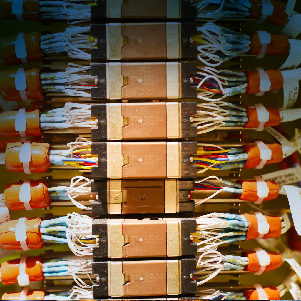Gros plan de fils électriques colorés connectés à des bornes avec des capuchons orange et des composants étiquetés, disposés en rangées verticales à l'intérieur d'un panneau électrique industriel.