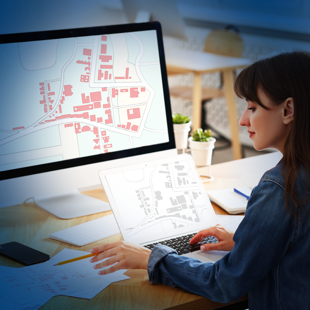 Une femme assise à un bureau analyse des cartes affichées sur un écran d'ordinateur et un ordinateur portable. Les cartes montrent le tracé des bâtiments et des rues en rouge et gris. L'espace de travail est aménagé avec des plantes et des fournitures de bureau.