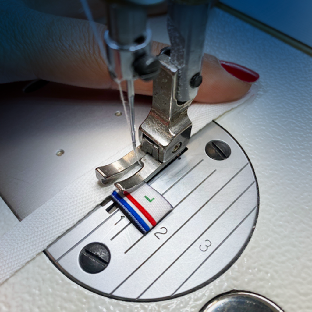 Gros plan d'une machine à coudre cousant un tissu blanc orné d'une étiquette rayée colorée. Une main, vernis à ongles rouge, guide le tissu sous le pied-de-biche.