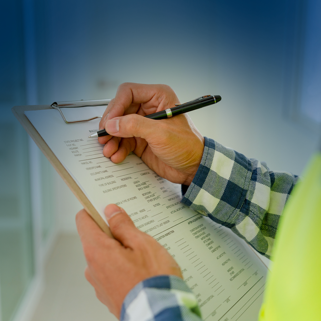 Une personne portant une chemise à carreaux et un gilet de sécurité remplit un formulaire sur un presse-papiers avec un stylo, effectuant probablement une inspection ou une enquête dans un environnement intérieur lumineux.