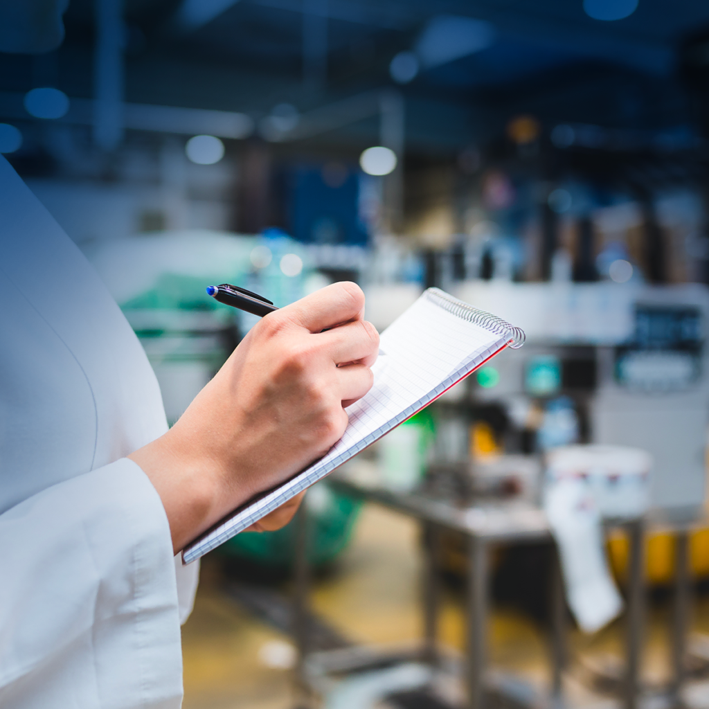 Une personne en blouse blanche tient un stylo et écrit sur un bloc-notes dans un environnement industriel ou de laboratoire avec des équipements et des machines flous en arrière-plan.