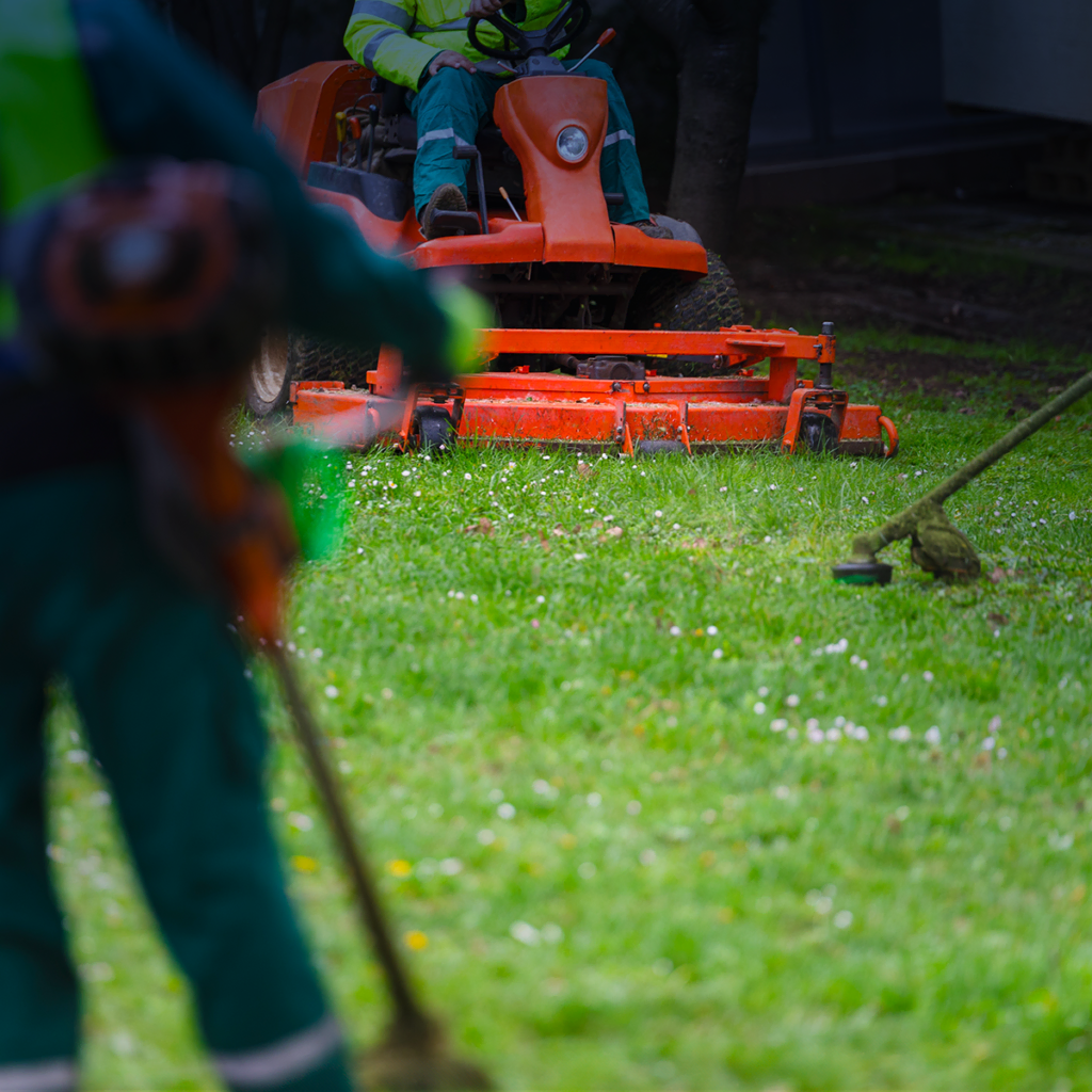 Deux ouvriers en uniformes verts entretiennent une pelouse herbeuse ; l'un conduit une tondeuse à gazon orange, tandis que l'autre utilise un coupe-bordures au premier plan.