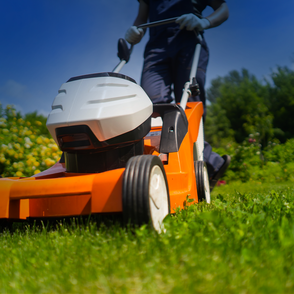 Une personne tond une pelouse verte avec une tondeuse orange dans un jardin, portant des gants. L'arrière-plan montre des buissons et des arbres sous un ciel bleu.