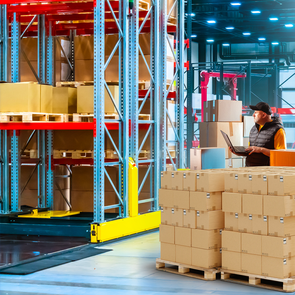 Dans un entrepôt, un ouvrier se tient devant des cartons empilés, utilisant un ordinateur portable. De hautes étagères remplies de cartons apparaissent en arrière-plan, sous un éclairage industriel intense.