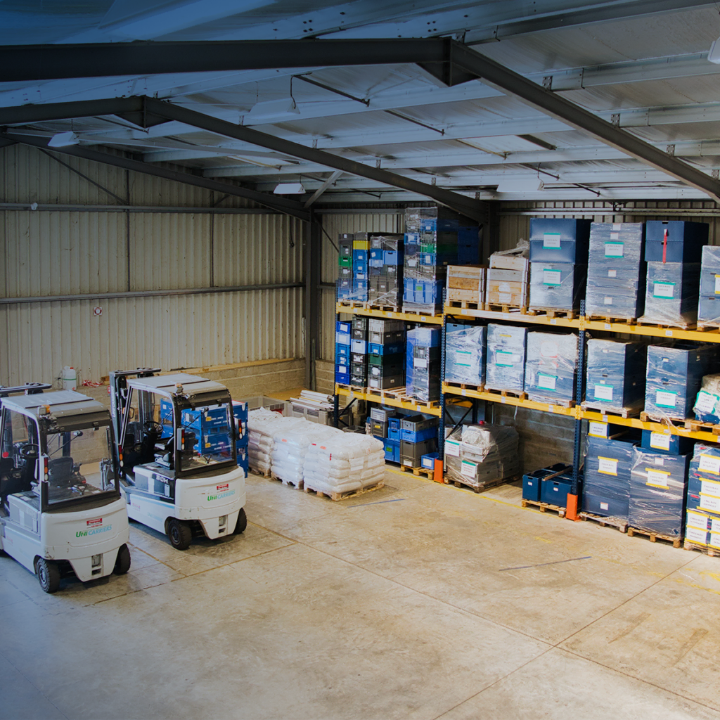 Un intérieur d'entrepôt avec deux chariots élévateurs stationnés à gauche et des étagères remplies de cartons et de palettes de marchandises à droite. Le sol est propre, l'espace est organisé et lumineux.