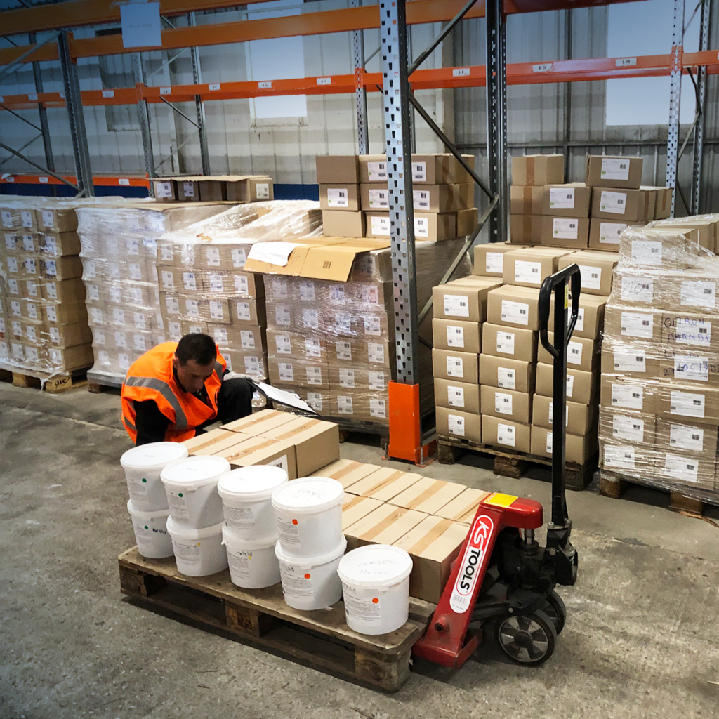 Un employé d'entrepôt portant un gilet de sécurité orange s'accroupit pour vérifier ou étiqueter des boîtes empilées sur une palette, entourée de palettes de boîtes en carton et de grands bacs en plastique blanc, avec des étagères en arrière-plan.