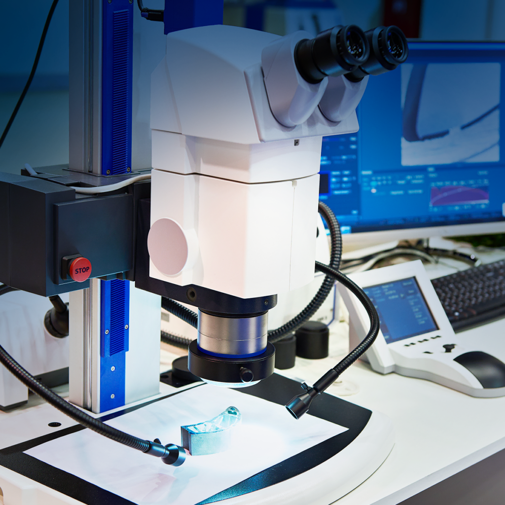 Un microscope de laboratoire moderne est posé sur un poste de travail, focalisé sur un petit objet métallique. À proximité, des écrans d'ordinateur affichent des images agrandies, et des équipements scientifiques entourent le microscope.