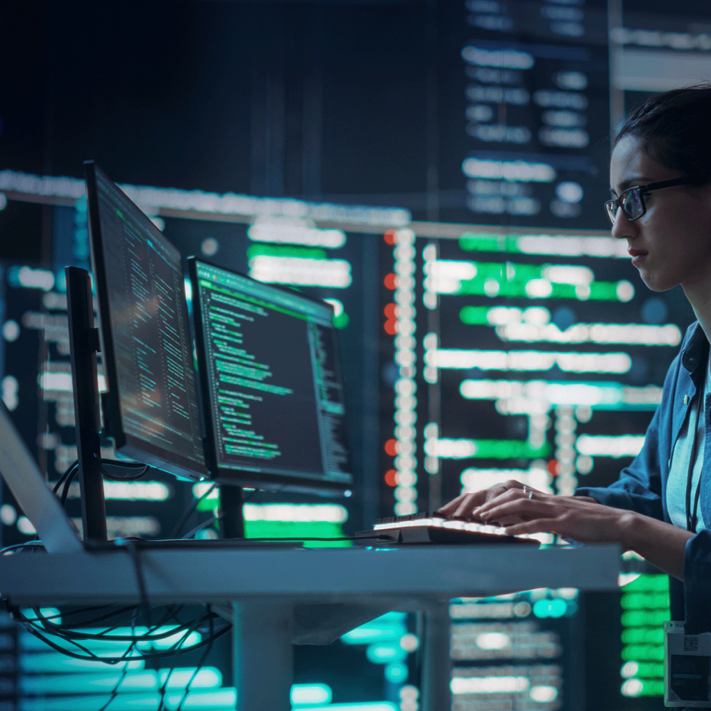 Une personne portant des lunettes travaille à un bureau avec plusieurs écrans d'ordinateur affichant du code, dans une pièce sombre avec de grands écrans numériques et des visualisations de données en arrière-plan.