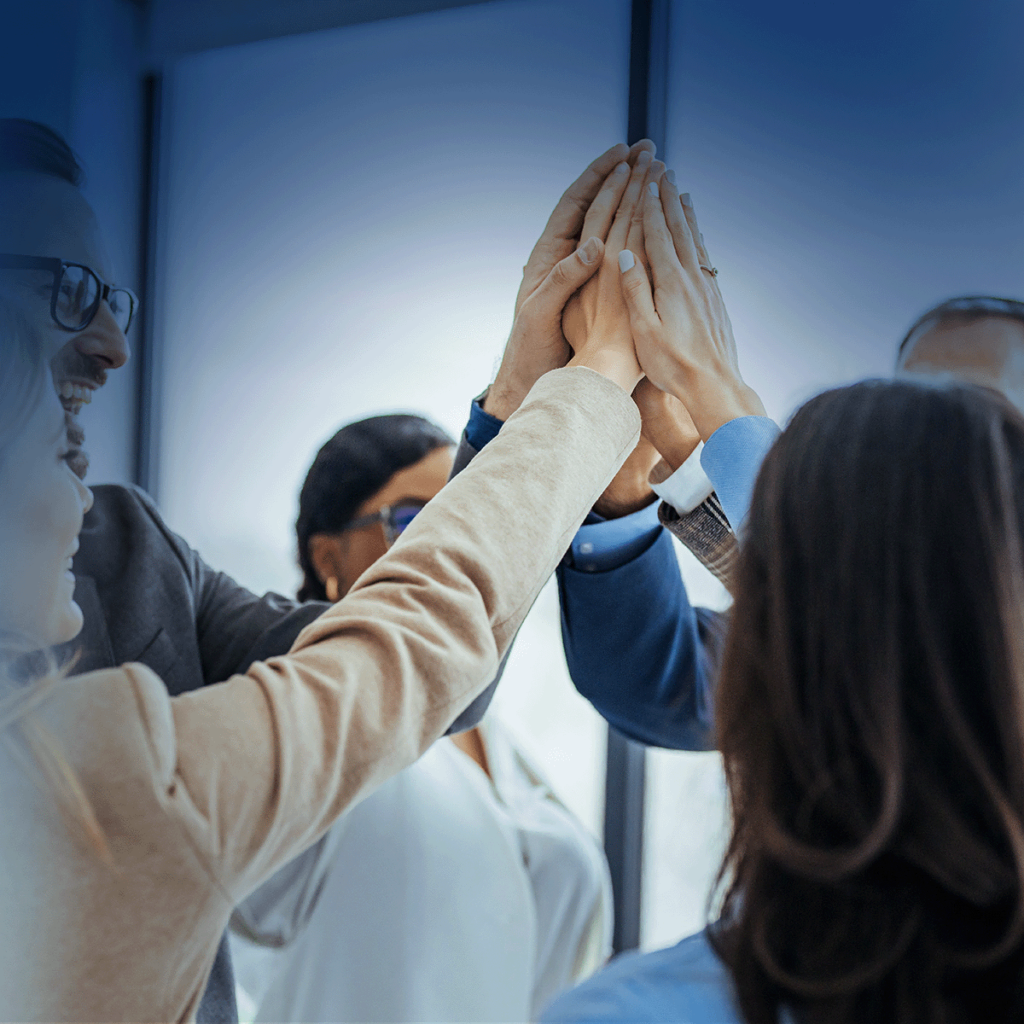 Un groupe de professionnels se tient en cercle, souriant et joignant les mains dans un geste de « high five », célébrant le travail d'équipe dans un environnement de bureau.