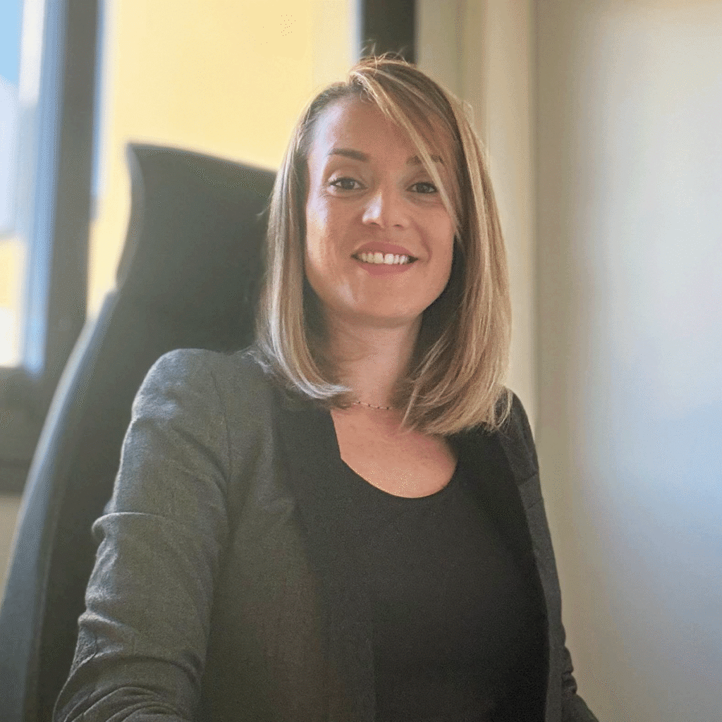 Une femme aux cheveux blonds raides et mi-longs, portant un blazer foncé sur un haut noir, est assise et sourit dans un bureau avec une fenêtre en arrière-plan.