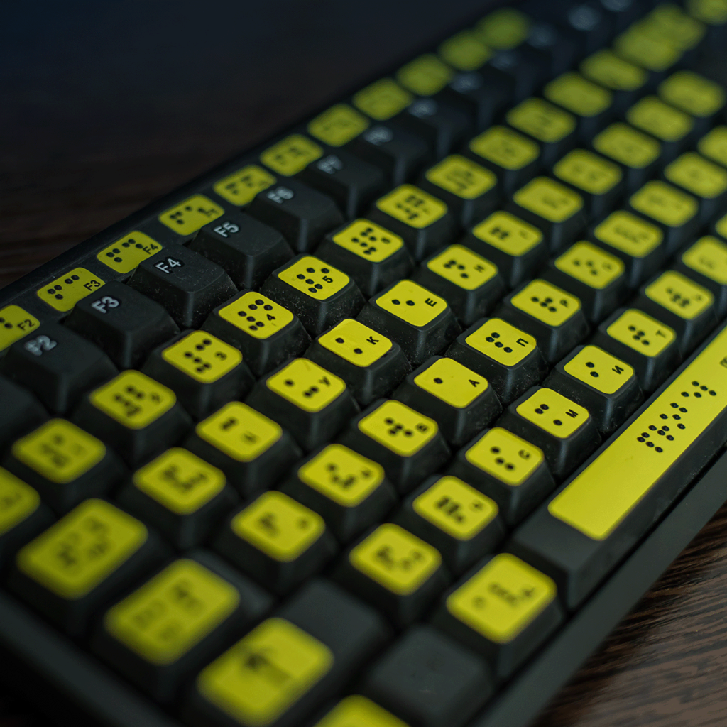 Un clavier d'ordinateur avec des touches jaunes affichant à la fois des lettres imprimées et des symboles Braille, conçu pour les utilisateurs malvoyants, repose sur une surface en bois sombre.