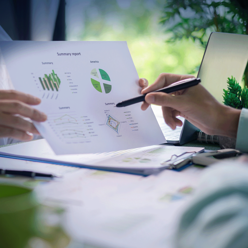 Deux personnes discutent d'un rapport de synthèse imprimé, accompagné de tableaux et de graphiques, en pointant les données avec un stylo. Des papiers et un ordinateur portable sont posés sur le bureau, et des plantes apparaissent en arrière-plan.