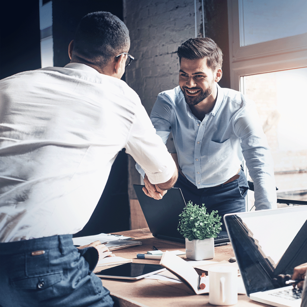 Deux hommes en tenue professionnelle souriant et se serrant la main à travers un bureau dans un bureau moderne, avec des ordinateurs portables, des papiers et une plante en pot visibles sur la table.