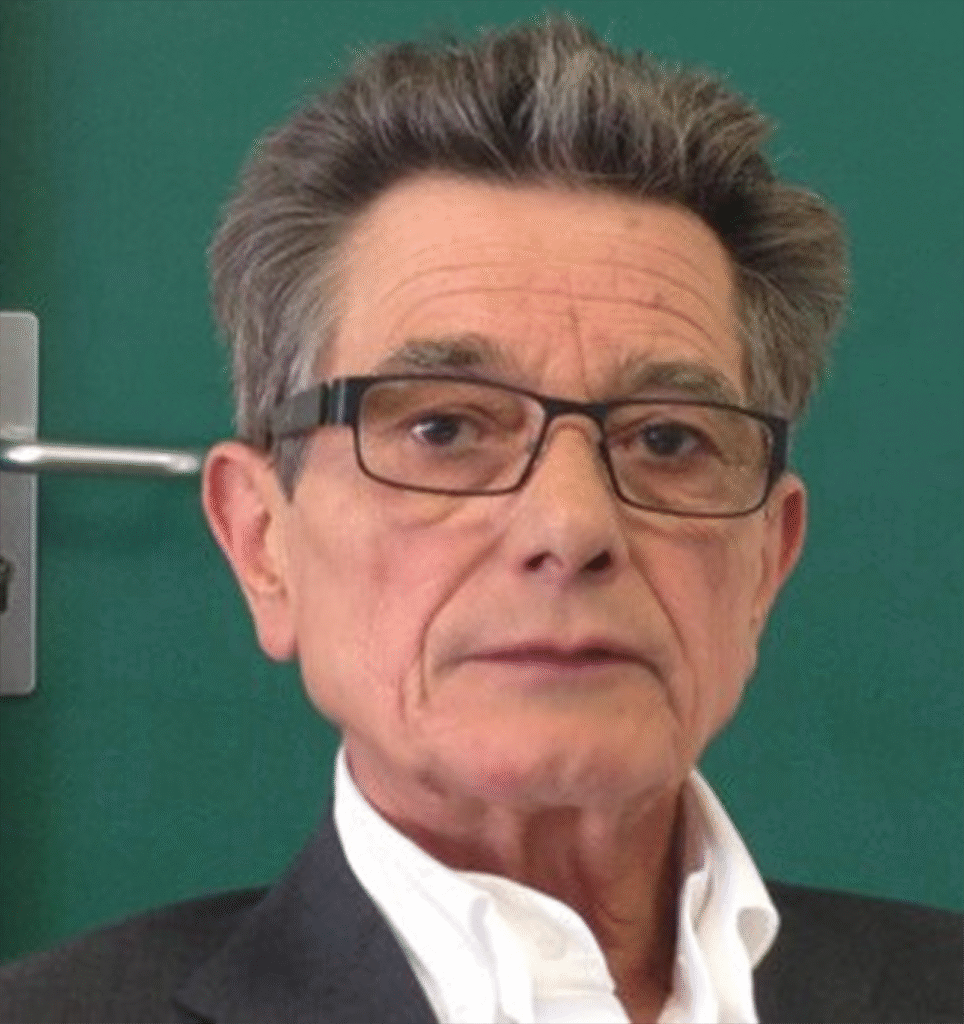 Un homme âgé aux cheveux gris courts et aux lunettes, vêtu d'une chemise blanche et d'une veste foncée, est assis devant un fond vert.