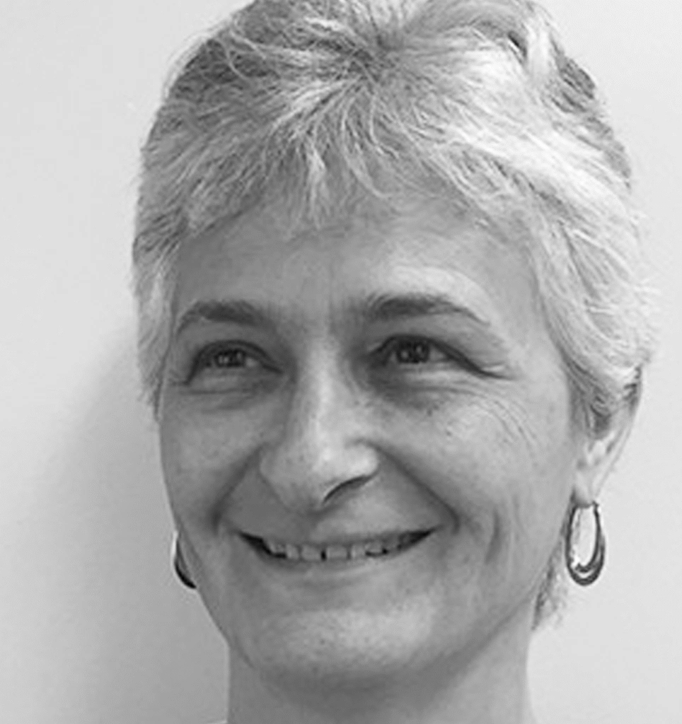 Photographie en noir et blanc d'une personne souriante aux cheveux courts et clairs, portant des boucles d'oreilles créoles, tournée légèrement vers la droite sur un fond uni.