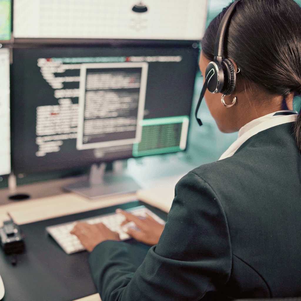 Une personne portant un casque et un costume travaille à un bureau avec plusieurs écrans affichant du code et des données dans un environnement de bureau moderne.