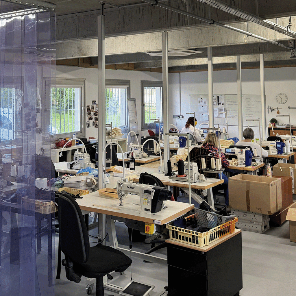 Plusieurs personnes travaillent à des machines à coudre dans un atelier industriel bien éclairé, avec de grandes fenêtres, des tables, des cartons et des fournitures visibles. Un rideau transparent pend à l'entrée.