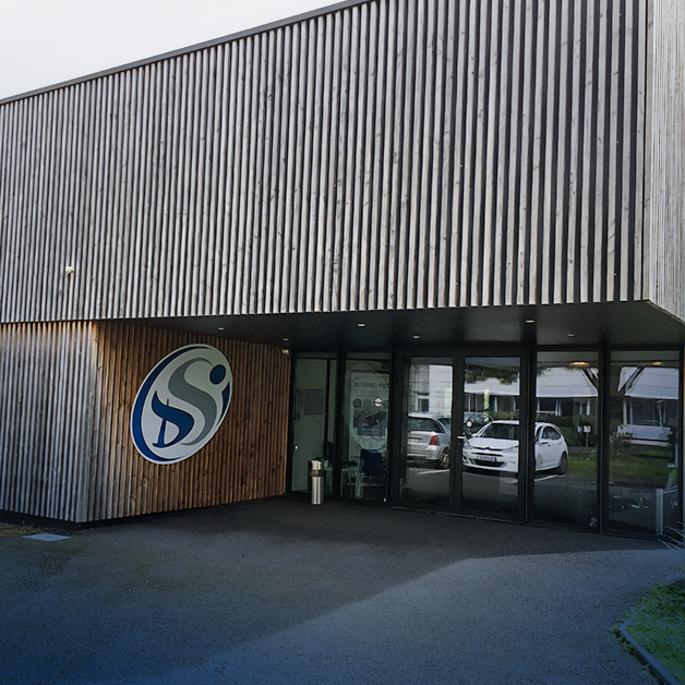 Un bâtiment moderne avec une façade en lattes de bois verticales, orné d'un grand logo circulaire bleu et blanc à l'entrée. Des portes vitrées mènent à l'intérieur, et les voitures garées sont visibles à travers les fenêtres.