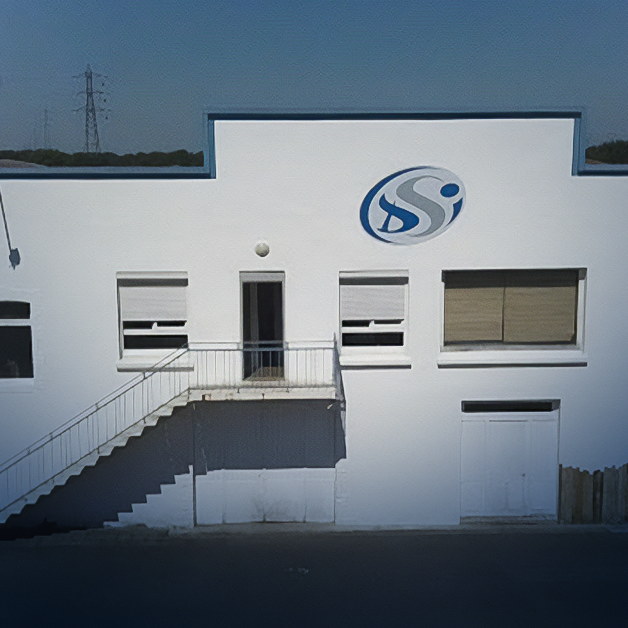 Un bâtiment industriel blanc avec un logo bleu et blanc au-dessus de l'entrée, deux fenêtres aux stores fermés et un escalier extérieur menant à une porte. Le ciel est dégagé et des lignes électriques sont visibles en arrière-plan.