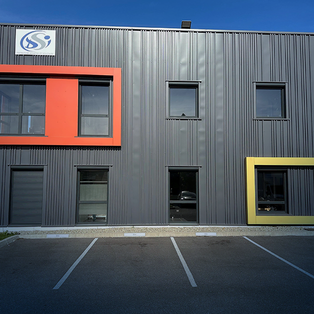 Un bâtiment industriel moderne avec un revêtement métallique gris, quatre fenêtres encadrées de cadres rouges et jaunes, une enseigne d'entreprise en haut à gauche et des places de parking vides devant.