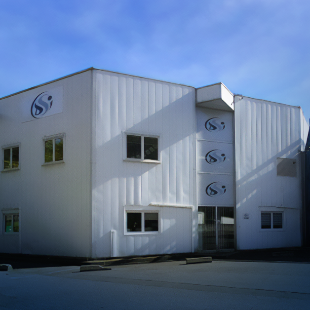Un bâtiment industriel blanc de deux étages avec plusieurs petites fenêtres et plusieurs logos noirs représentant un design abstrait en S à l'extérieur ; le ciel est bleu avec des nuages doux.
