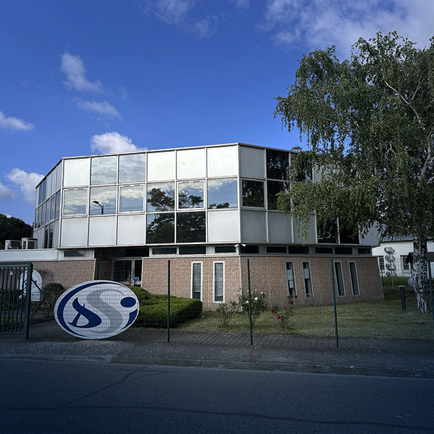 Un immeuble de bureaux moderne de deux étages, doté de grandes fenêtres réfléchissantes et d'un soubassement en briques, se dresse derrière une clôture grillagée par une journée ensoleillée. Un panneau circulaire bleu et blanc au design abstrait est installé près de l'entrée.