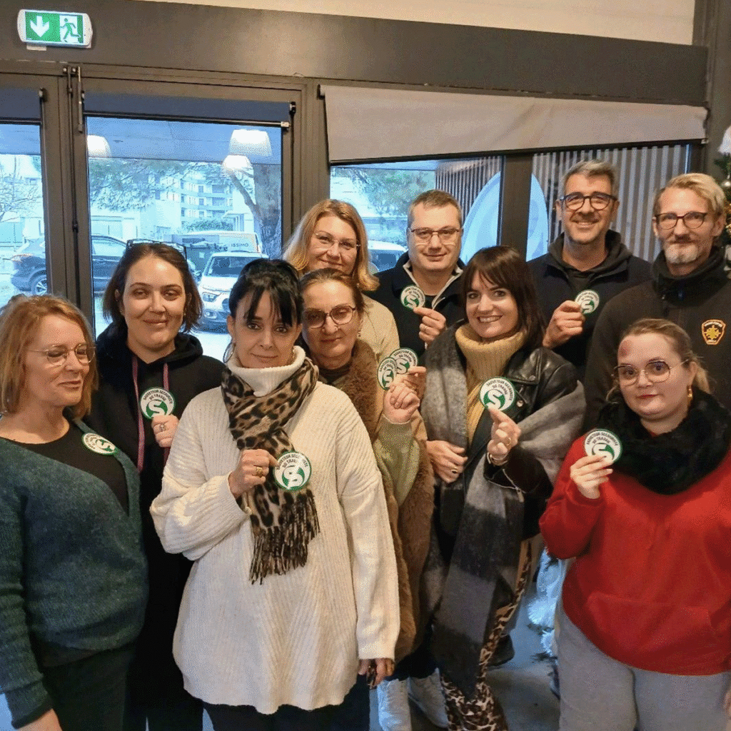 Un groupe de dix adultes se tient à l'intérieur, souriant et tenant des badges ronds verts et blancs. Derrière eux, de grandes portes et fenêtres vitrées laissent entrevoir des bâtiments et des arbres.