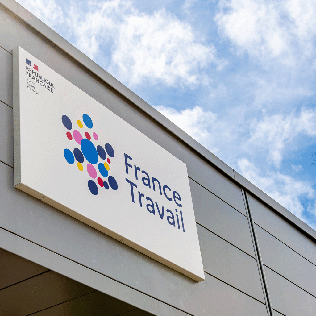 Un panneau France Travail à pois colorés est installé sur un bâtiment gris, sous un ciel bleu partiellement nuageux. Le logo de la République française est visible dans le coin supérieur gauche du panneau.