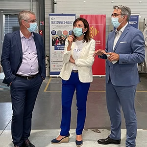 Trois personnes portant des masques et une tenue professionnelle se tiennent à l'intérieur et discutent. Derrière elles, des affiches d'information en français, dont une pour « Cap sur l'Emploi », et des affiches de matériel industriel ou de bureau.