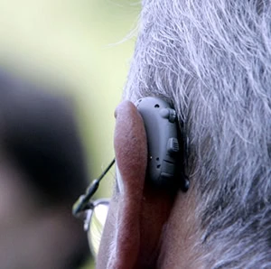 Gros plan sur l'oreille d'une personne âgée équipée d'un appareil auditif gris « Dispositif médical pour malentendants », montrant des cheveux gris et une partie de l'oreille. L'arrière-plan est flou.