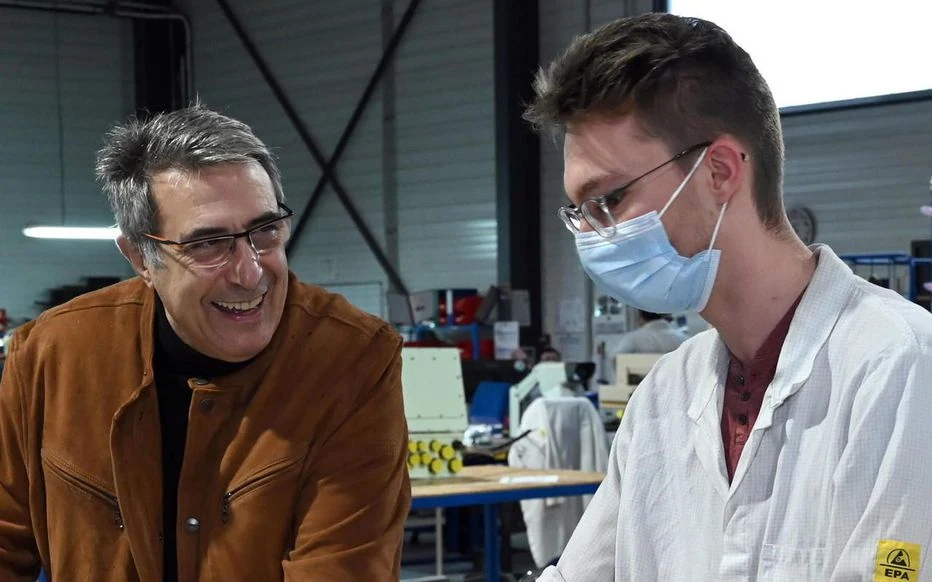 Deux hommes discutent et sourient dans un atelier toulousain ; l'un porte une veste marron et des lunettes, l'autre une blouse blanche, des lunettes et un masque. Le matériel industriel et les tables bleues reflètent le travail du groupe DSI dans le secteur économique et social.