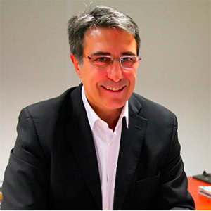 Jean-Louis RIBES, aux cheveux gris courts et portant des lunettes, sourit dans une veste de costume noire et une chemise blanche alors qu'il est assis à un bureau lors d'une interview dans un cadre intérieur bien éclairé.