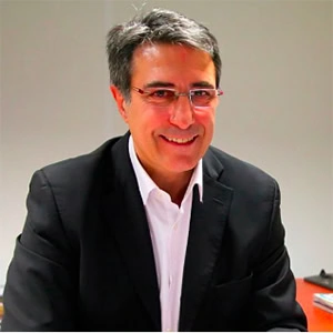 Jean-Louis RIBES, aux cheveux gris courts et portant des lunettes, sourit dans une veste de costume noire et une chemise blanche alors qu'il est assis à un bureau lors d'une interview dans un cadre intérieur bien éclairé.