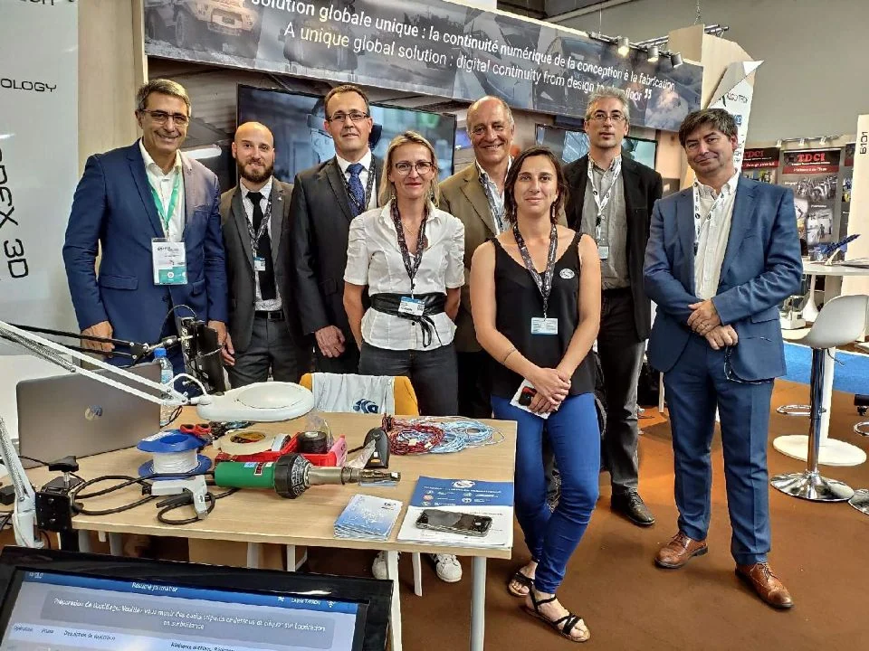 Un groupe de neuf professionnels pose et sourit pour une photo sur le stand de DSI. La table devant présente divers appareils et brochures technologiques, avec en arrière-plan les bannières du Salon du Bourget SIAE 2019.
