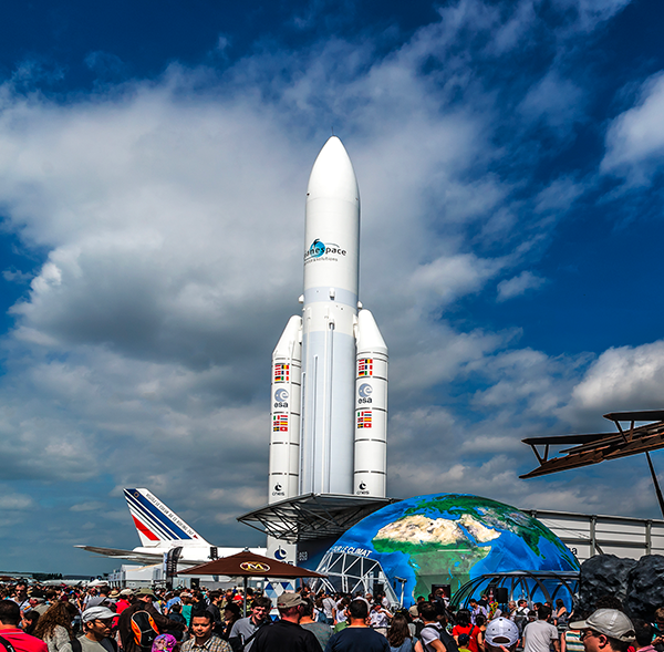 Une foule se rassemble autour d'une grande fusée blanche exposée en plein air, avec une maquette de la Terre et un avion à proximité, sous un ciel bleu partiellement nuageux.