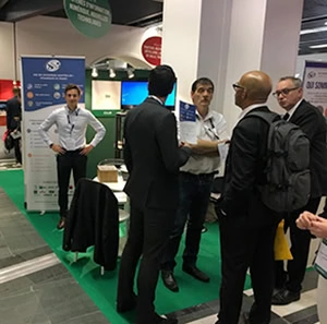 Un groupe d'hommes en tenue professionnelle discutent sur un stand du Salon du Handicap à Paris, avec des affiches et des présentoirs d'information DSI en arrière-plan sur une zone recouverte de moquette verte.